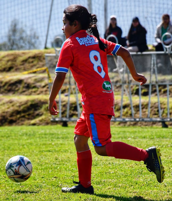 services-01 A girl in a red jersey playing soccer outdoors on grass with spectators in background.