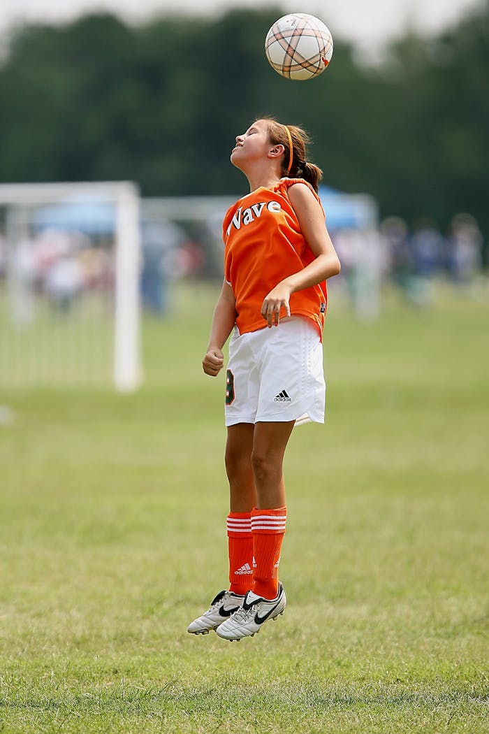 services-02 A young girl jumps to head a soccer ball during a game on a grassy field.